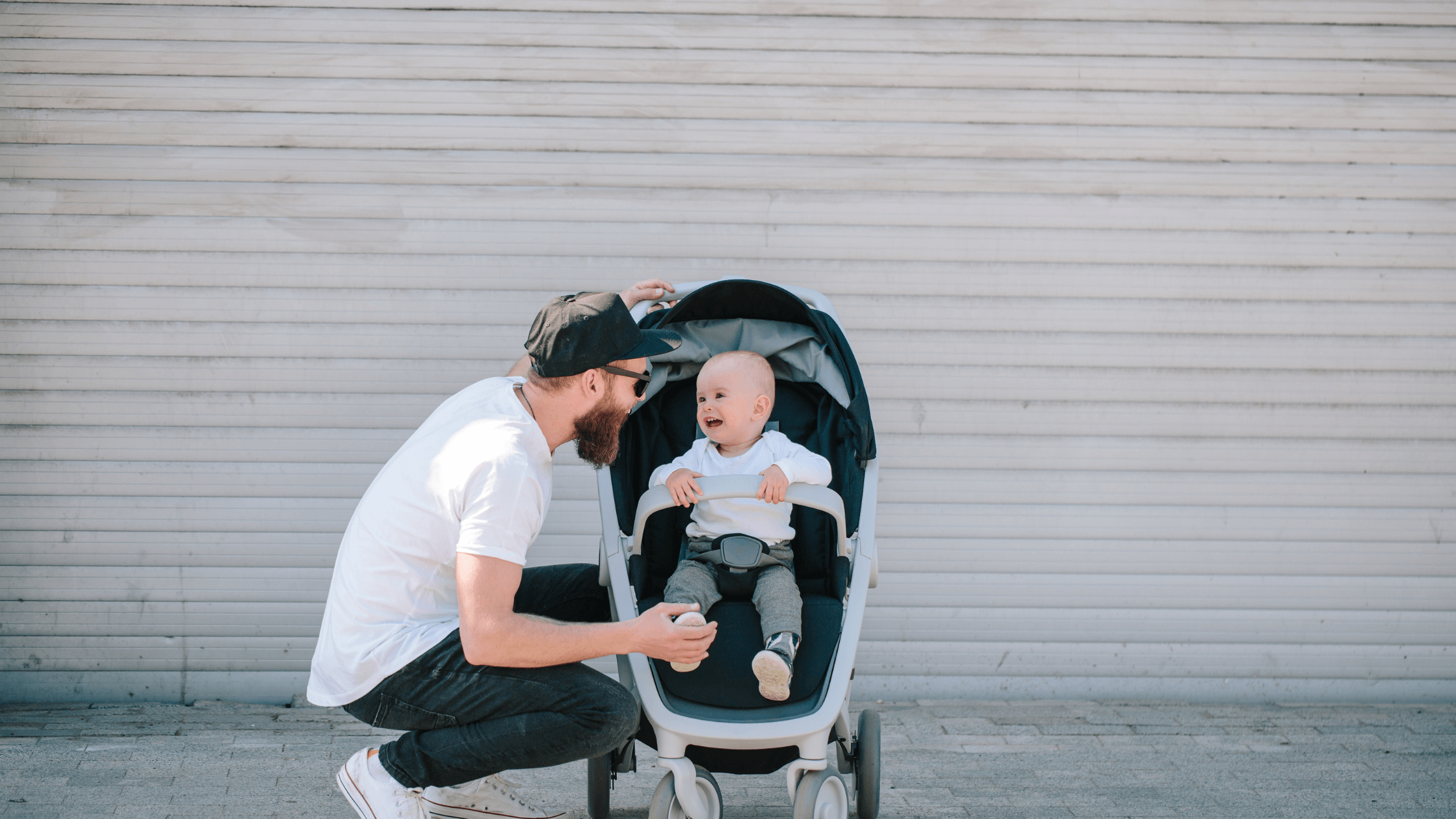A father with a beard and baseball cap crouching down to interact with a smiling baby in a stroller.