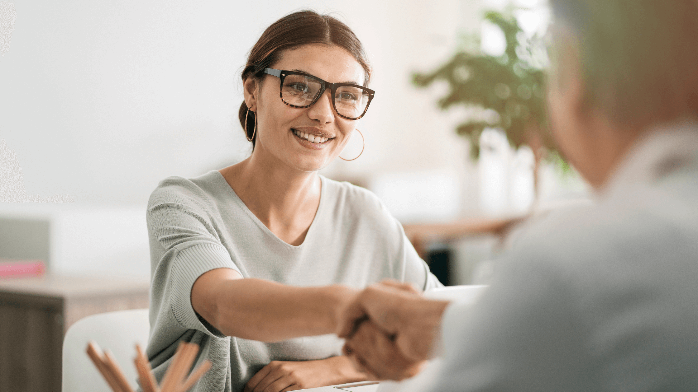 A woman with glasses shakes hands with a person across a table, symbolizing a successful consultation or agreement.