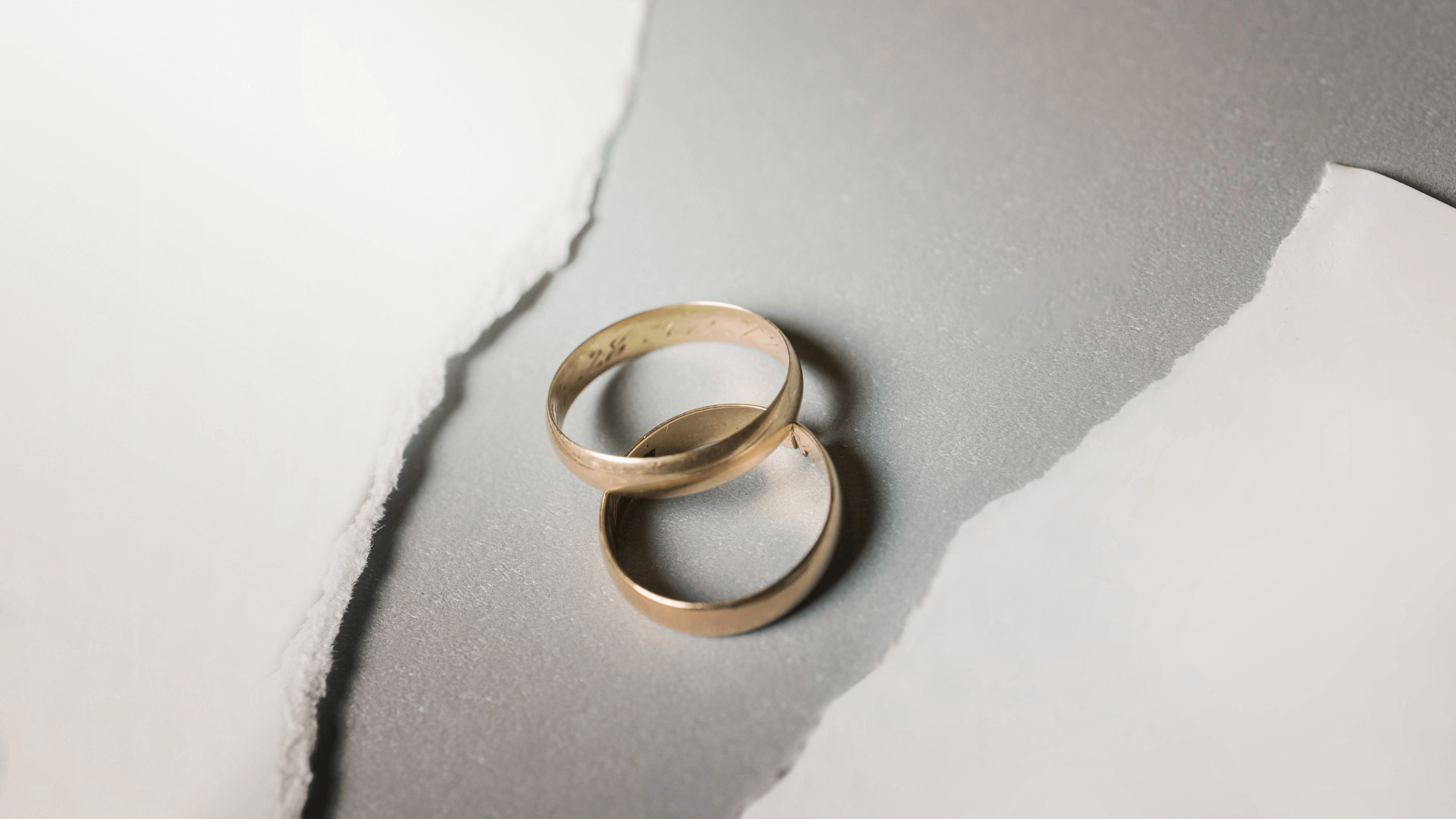 Two gold wedding rings are separated on a grey surface next to torn paper, symbolizing the end of a marriage and legal finalization.