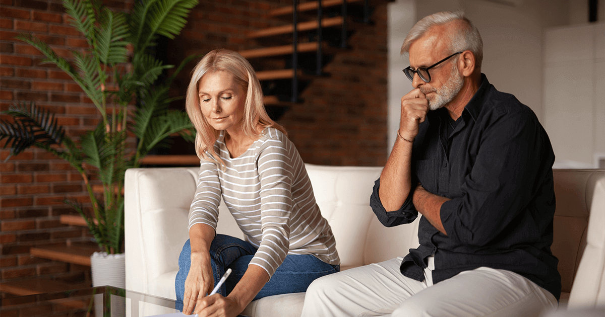 An image of an older man and woman sitting on a couch reviewing documents, with the woman writing and the man looking pensive, symbolizing a couple reviewing financial and separate property documents during a divorce consultation.